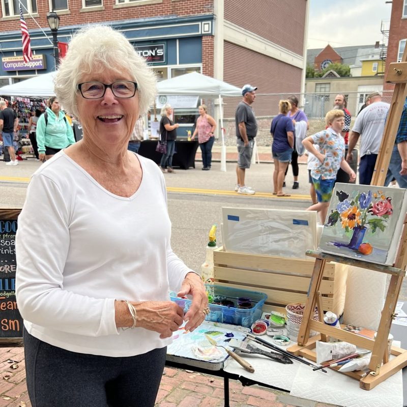 An artist smiles while holding paintbrushes, with a colorful painting and supplies on display. People walk by in the background.
