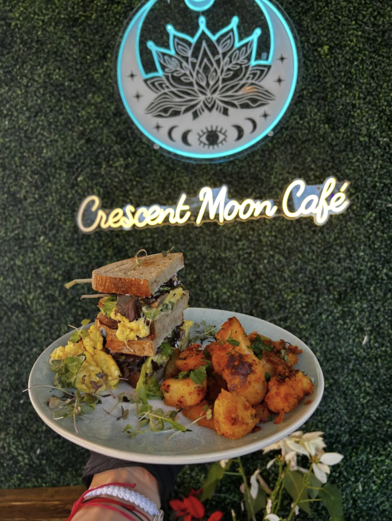 A hand holds a plate with a sandwich, scrambled eggs, and seasoned potatoes in front of a café sign.